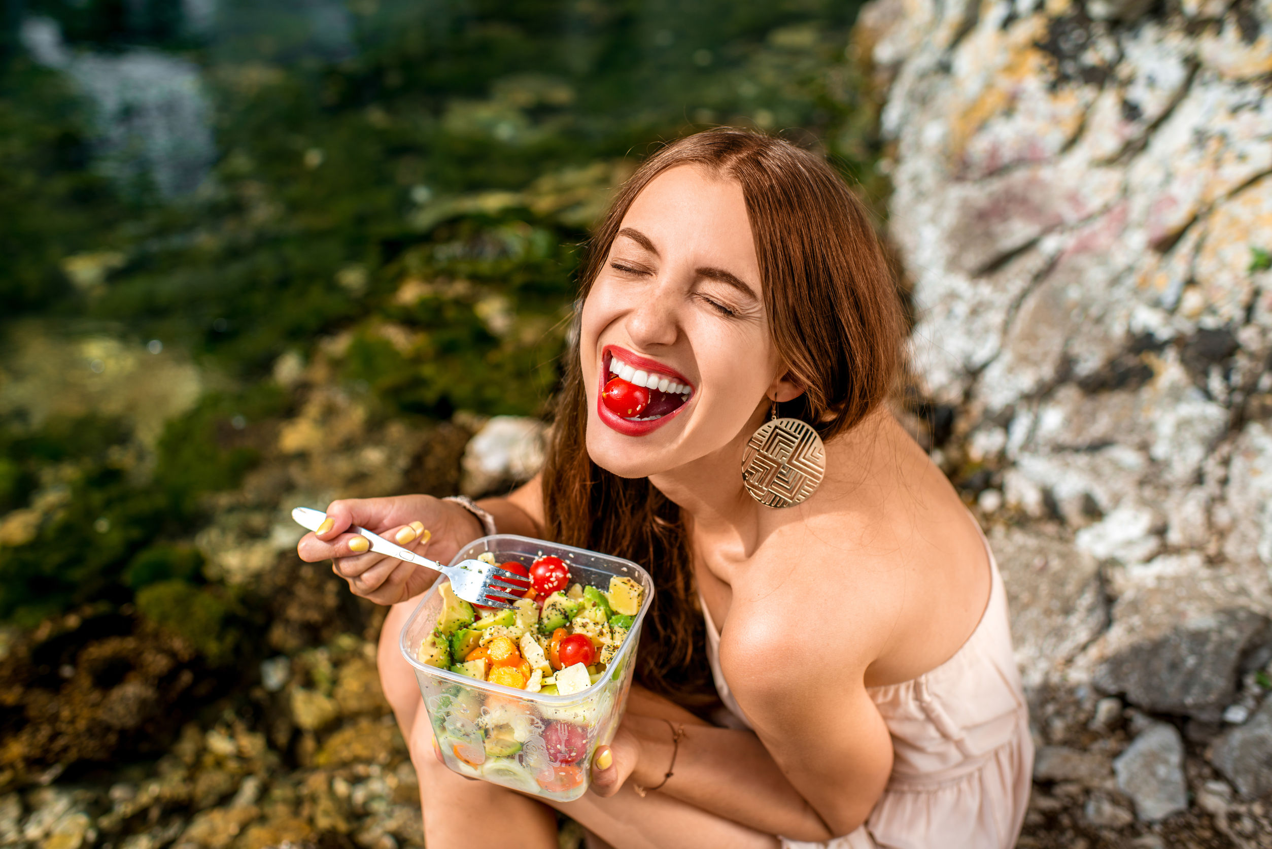 woman eating healthy salad from plastic container near the river, holistic health coach